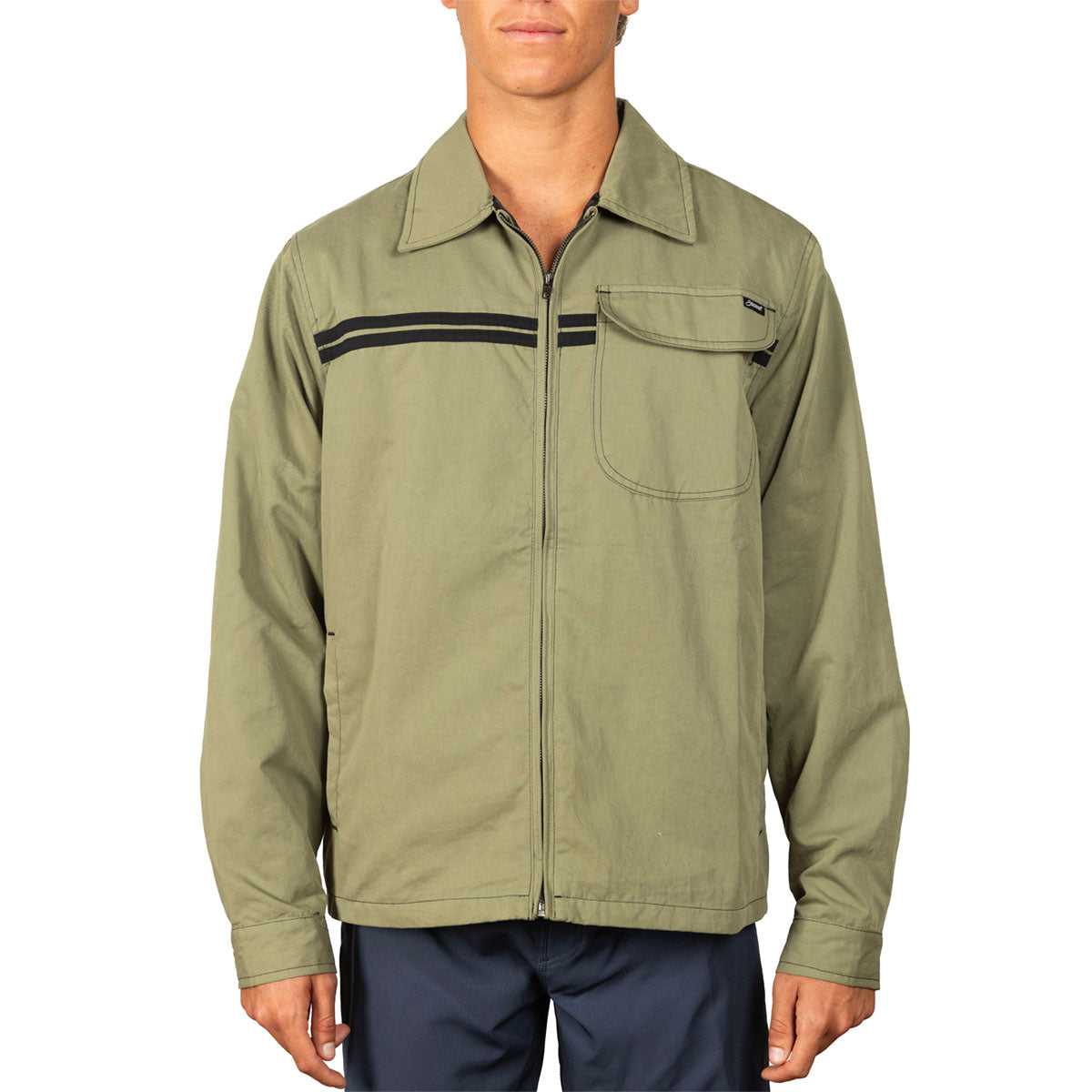 A man wearing an olive green zip-up work jacket with black horizontal stripes across the chest, paired with navy pants, against a plain white background.