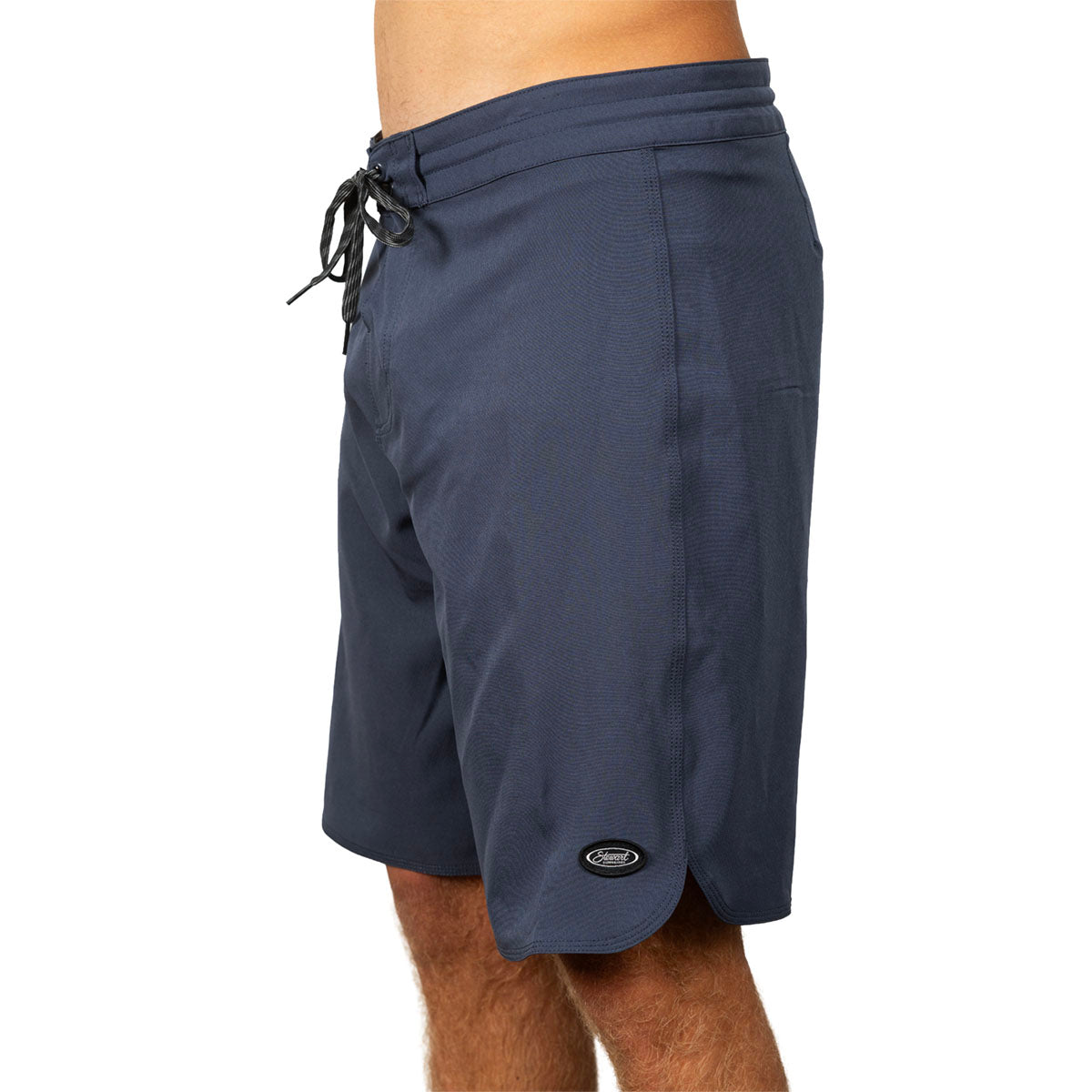 A side view of a person wearing navy blue board shorts with a black drawstring and a small oval logo patch near the hem, against a plain white background.