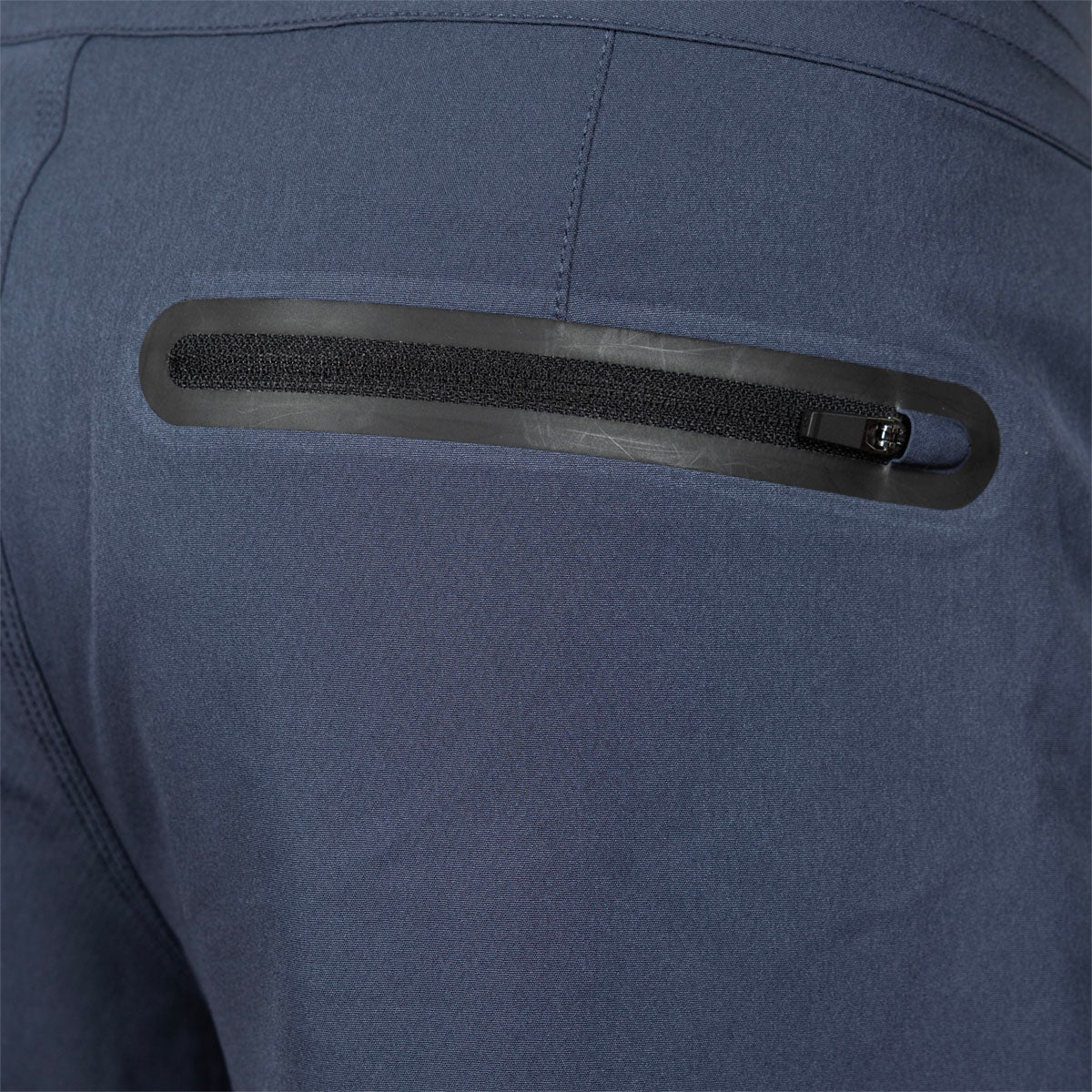 Close-up of a dark blue technical jacket showing a horizontal zippered pocket with a black zipper track and a small metal pull tab.