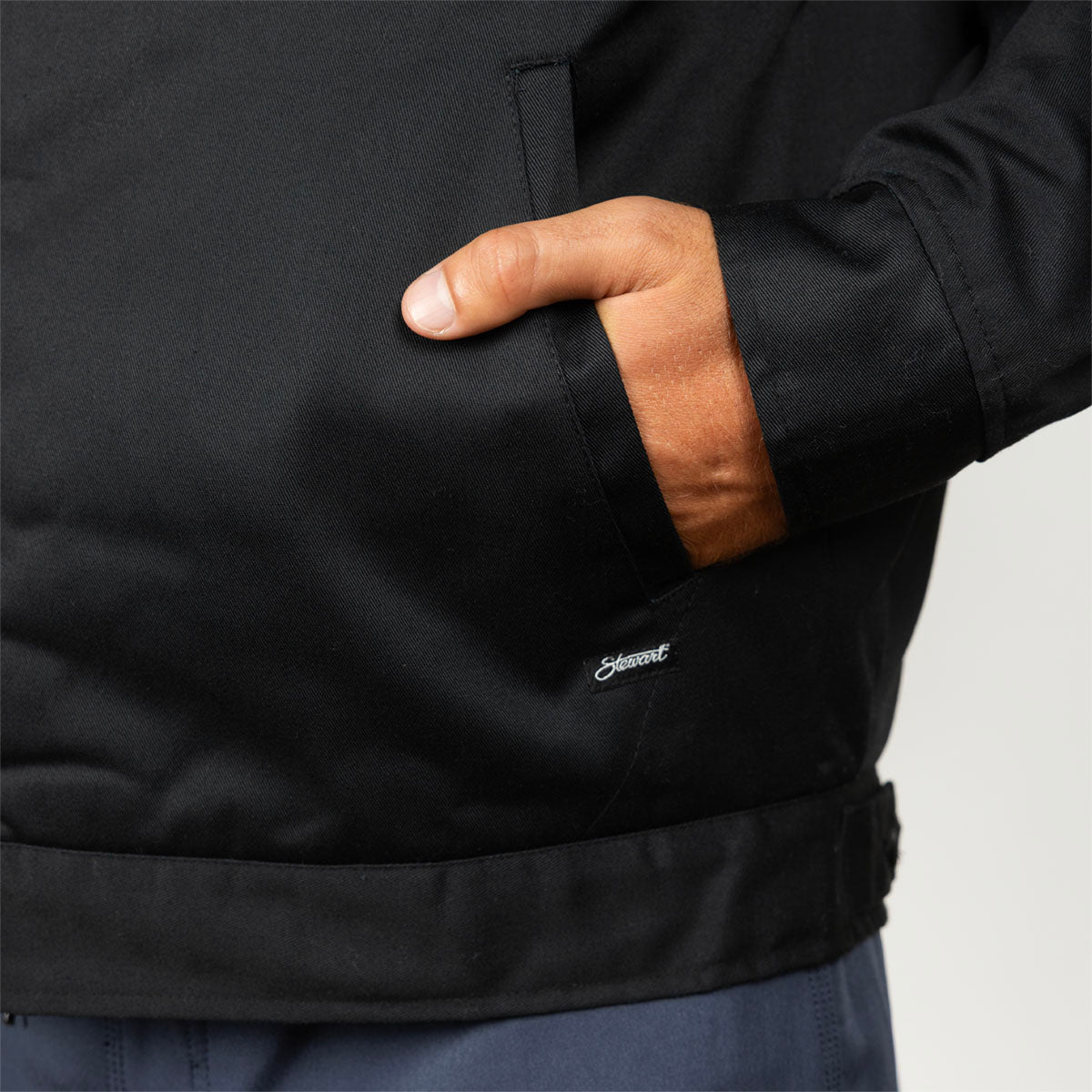 Close-up of a person wearing a black jacket, showing a hand in a side pocket and a small white embroidered Stewart logo near the lower front.