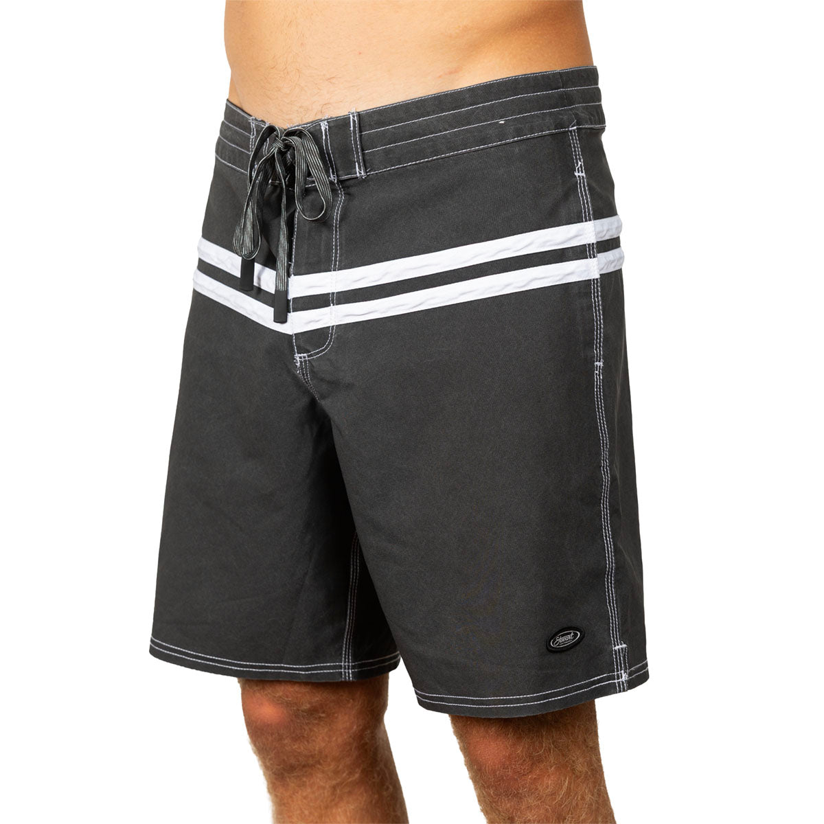 A close-up of a person wearing dark charcoal board shorts with two bold white horizontal stripes across the front waistband and white contrast stitching, shown against a plain white studio background.