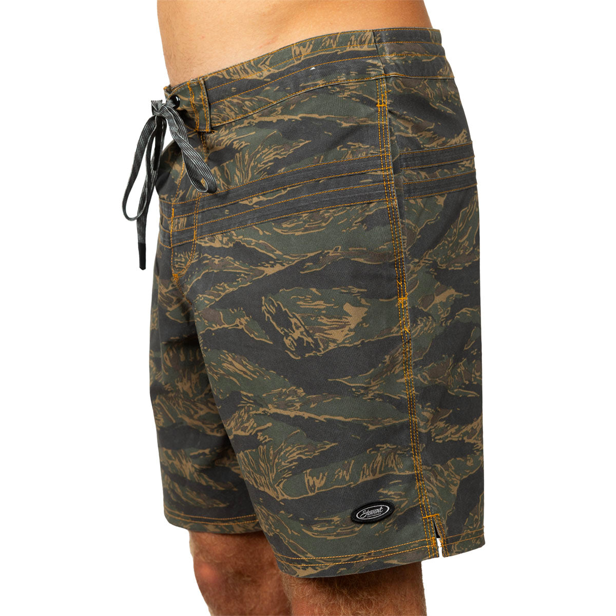 A close-up of a person wearing board shorts with an allover camouflage-style pattern in olive green, black, and tan, featuring a black drawstring and a small oval logo patch near the hem.