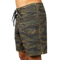 A close-up of a person wearing board shorts with an allover camouflage-style pattern in olive green, black, and tan, featuring a black drawstring and a small oval logo patch near the hem.