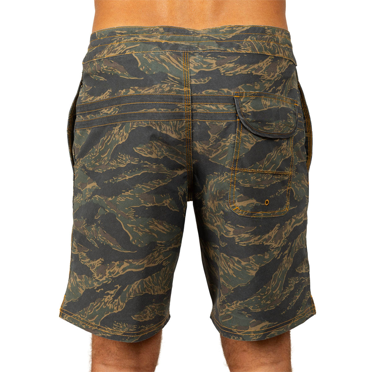 Rear view of a person wearing camouflage board shorts with a tiger-stripe pattern in olive green, black, and tan, on a white background.