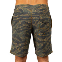 Rear view of a person wearing camouflage board shorts with a tiger-stripe pattern in olive green, black, and tan, on a white background.