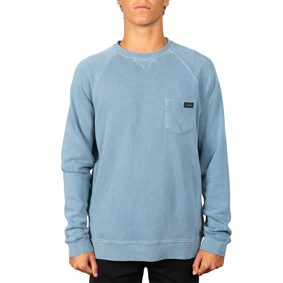 A man wearing a light blue long-sleeve crewneck sweatshirt with a chest pocket, shown from the neck down against a plain white background.