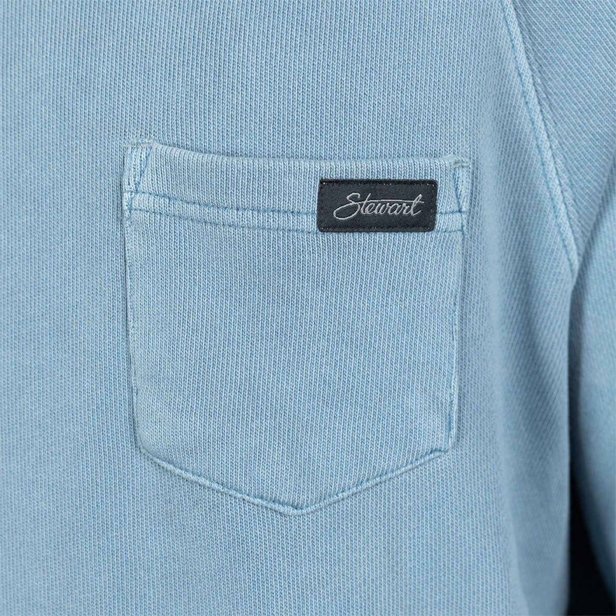 Close-up of a light blue textured fabric garment featuring a front chest pocket with a black rectangular Stewart brand tab.