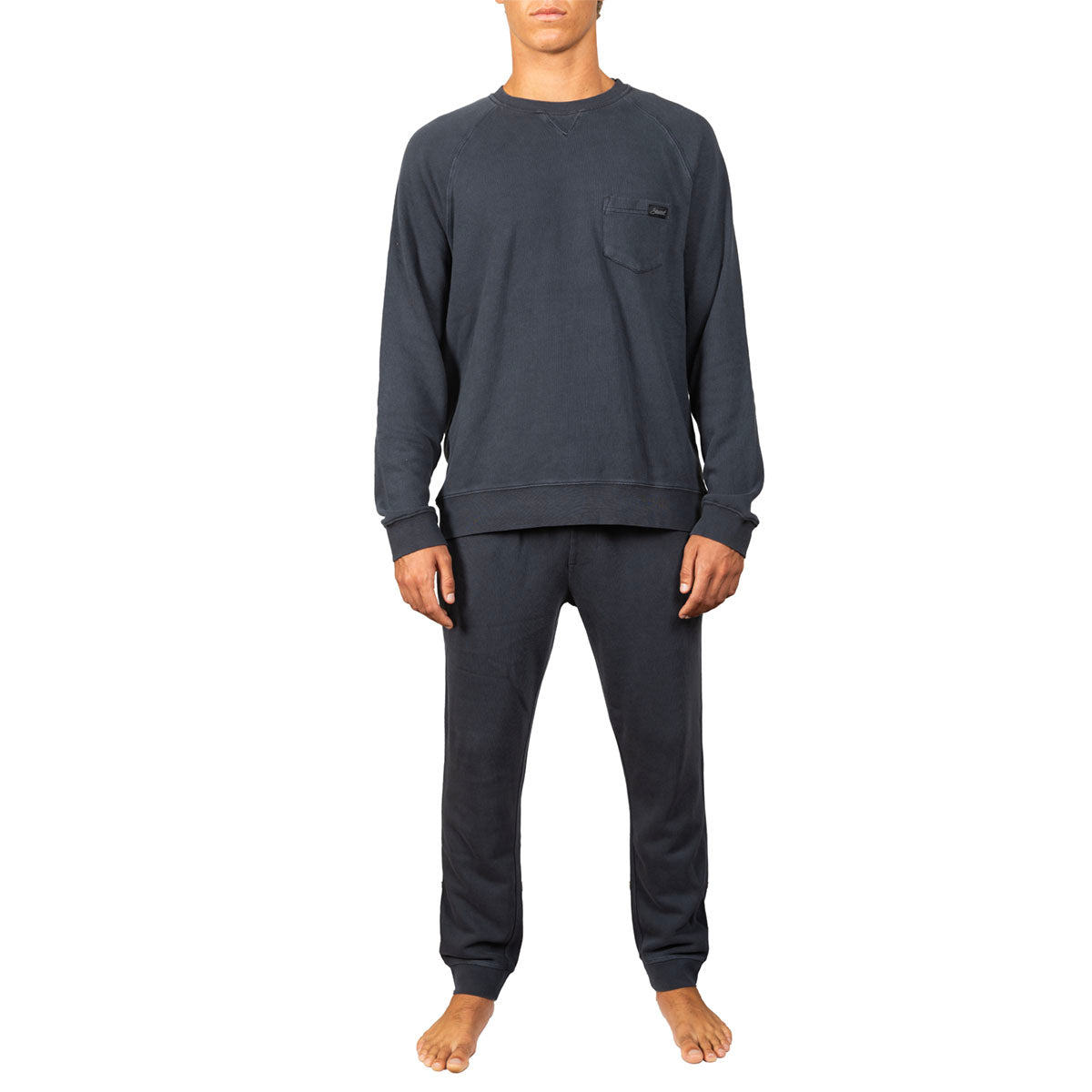 A man modeling a dark charcoal gray crewneck sweatshirt and matching sweatpants set against a plain white background.