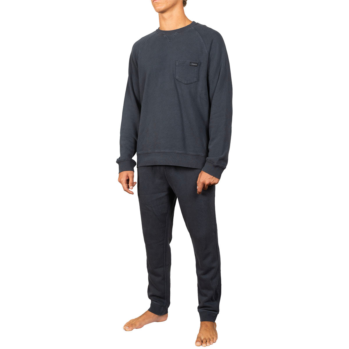 A man wearing a dark charcoal gray long-sleeve crewneck sweatshirt with a chest pocket and matching dark gray sweatpants, shown against a plain white background.