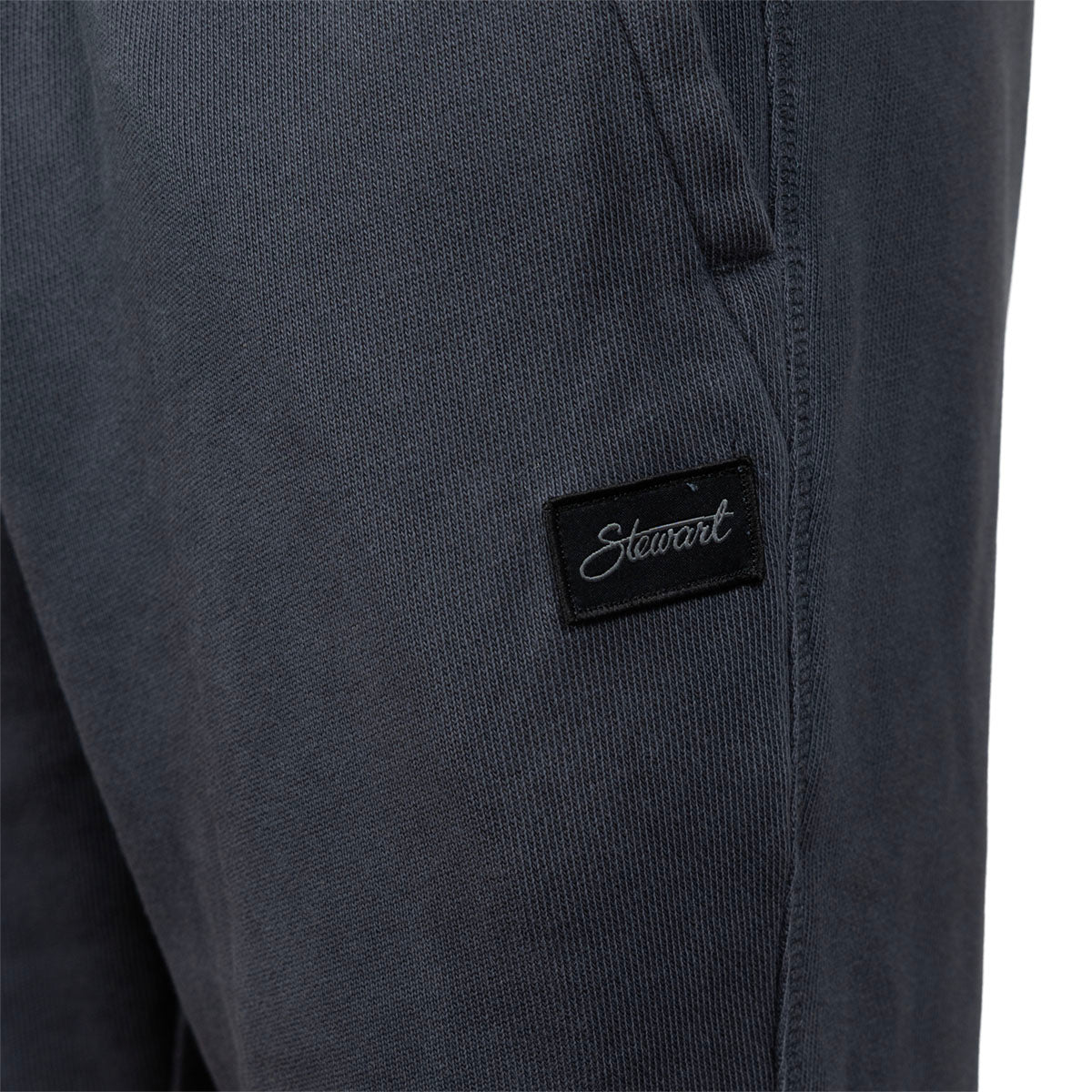 Close-up of dark charcoal gray fabric pants with a black rectangular patch on the leg featuring the white script brand name 'Stewart'.
