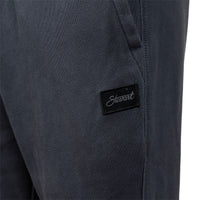 Close-up of dark charcoal gray fabric pants with a black rectangular patch on the leg featuring the white script brand name 'Stewart'.
