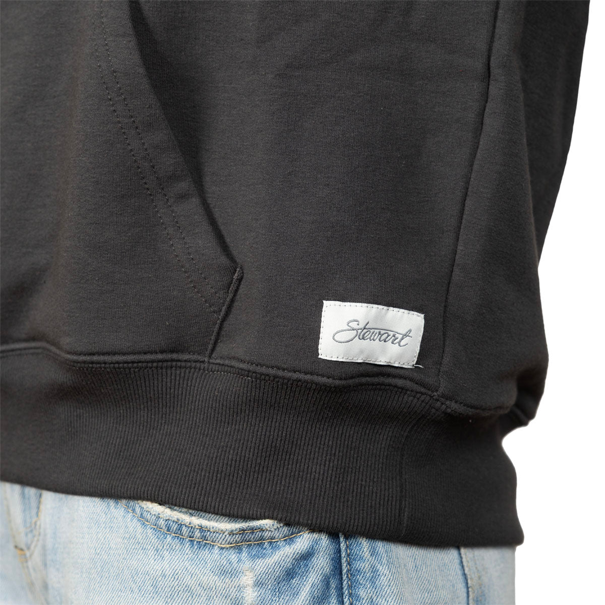 Close-up of a black sweatshirt worn with light blue jeans, showing a white rectangular brand patch with the script logo 'Stewart' near the lower right front hem.