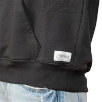 Close-up of a black sweatshirt worn with light blue jeans, showing a white rectangular brand patch with the script logo 'Stewart' near the lower right front hem.