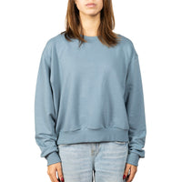 A woman wearing a dusty blue crewneck sweatshirt and light-wash jeans, shown from the neck down against a plain white background.