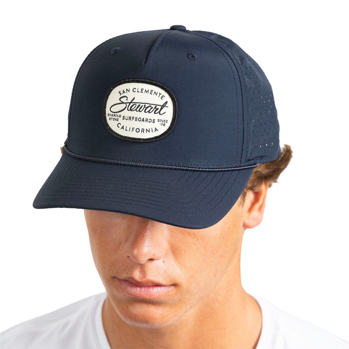 A man wearing a navy blue baseball cap with a circular embroidered patch on the front that reads 'San Clemente Stewart California'.