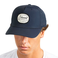 A man wearing a navy blue baseball cap with a circular embroidered patch on the front that reads 'San Clemente Stewart California'.