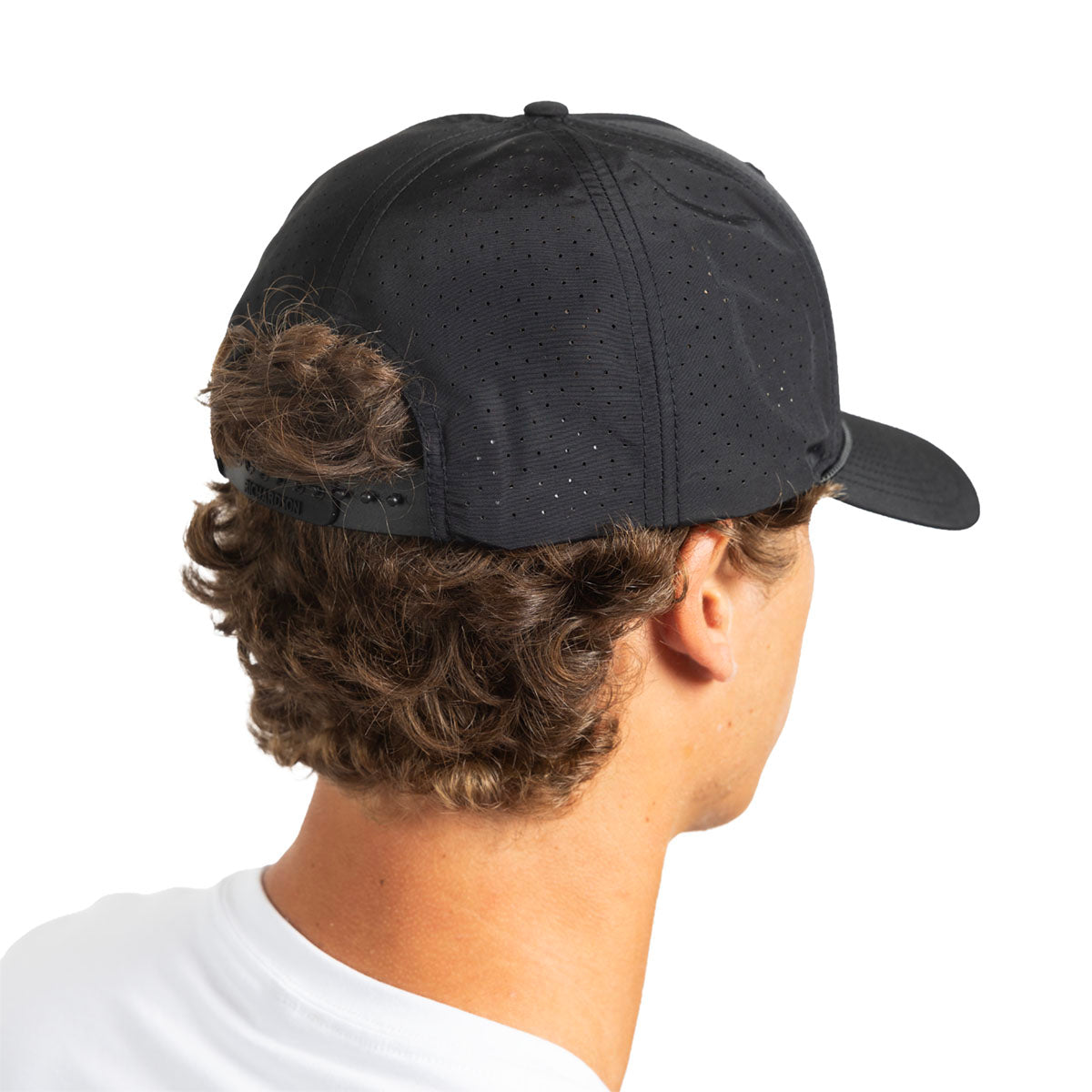 A man shown from the back wearing a black perforated baseball cap, with curly brown hair and a white t-shirt visible against a plain white background.