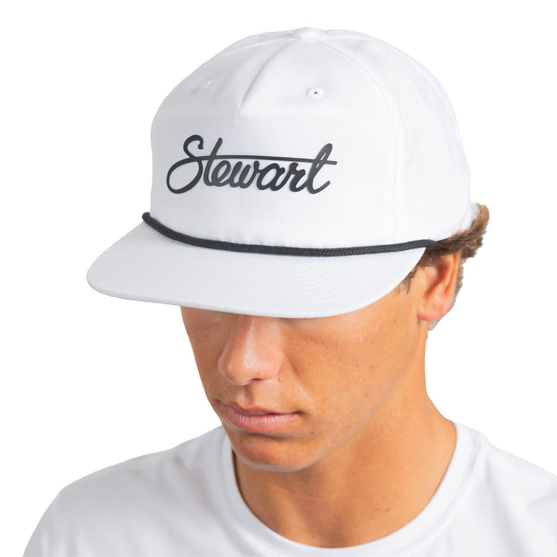 A man wearing a white baseball cap with 'Stewart' embroidered in black script across the front and a thin black horizontal stripe under the brim, photographed against a plain white background.