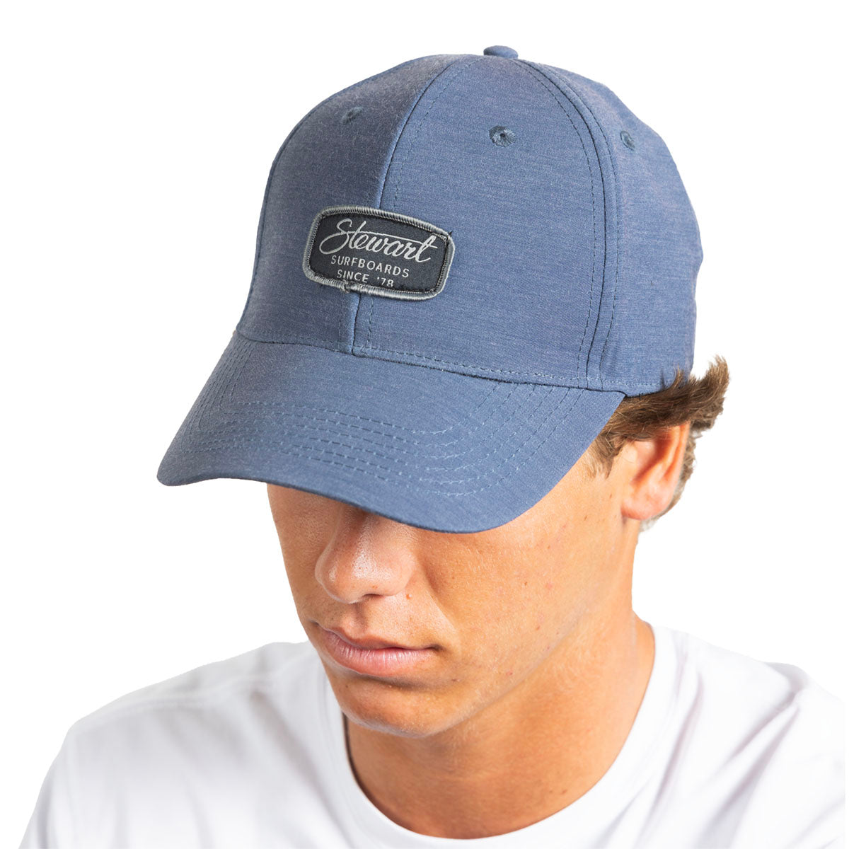 A man wearing a heathered slate blue baseball capwith a black rectangular patch on the front that reads 'Stewart Surfboards Since '78