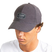 A man wearing a dark heather gray baseball cap with a black rectangular patch on the front that reads 'Stewart Surfboards Since '78