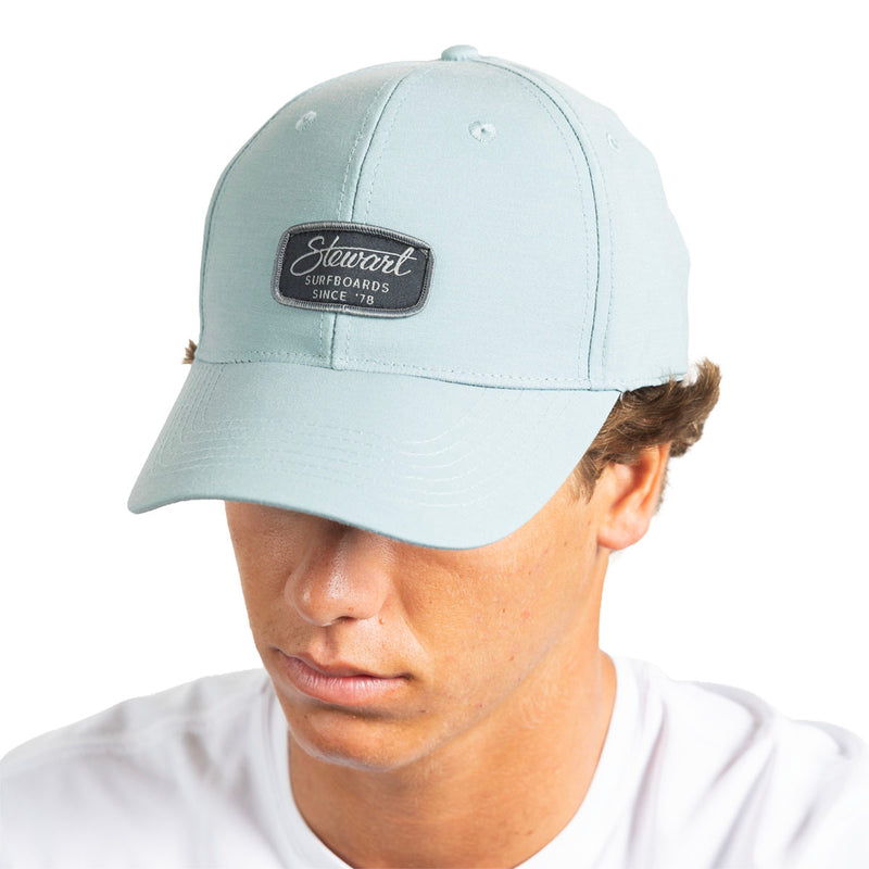 A man wearing a light blue baseball cap with a black rectangular patch on the front that reads 'Stewart Surfboards Since '78