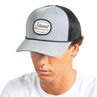 A man wearing a light gray and black trucker hat with a circular embroidered patch on the front that reads 'San Clemente Stewart California'.
