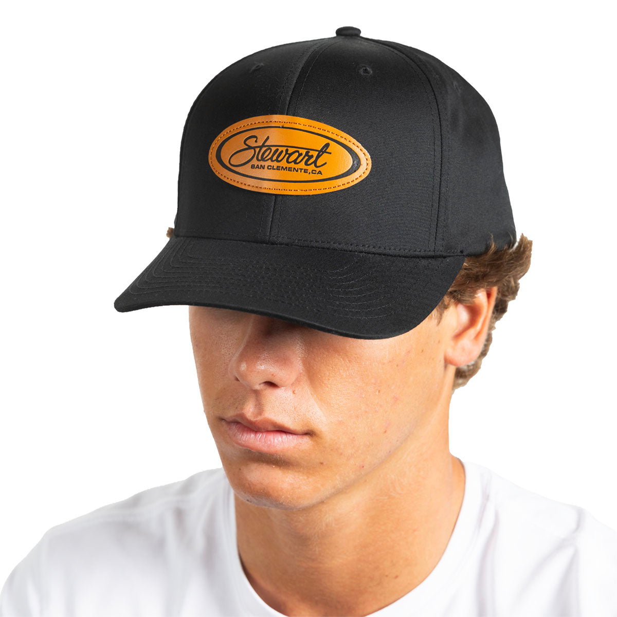 A man wearing a black baseball cap with an oval tan leather patch on the front that reads 'Stewart ' in black script, photographed against a plain white background.