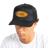 A man wearing a black trucker-style cap with a curved brim and a brown leather patch on the front that reads 'Stewart San Clemente'. The background is solid white.