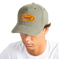 A man wearing an olive green baseball cap with an oval embroidered patch on the front that reads 'Stewart'