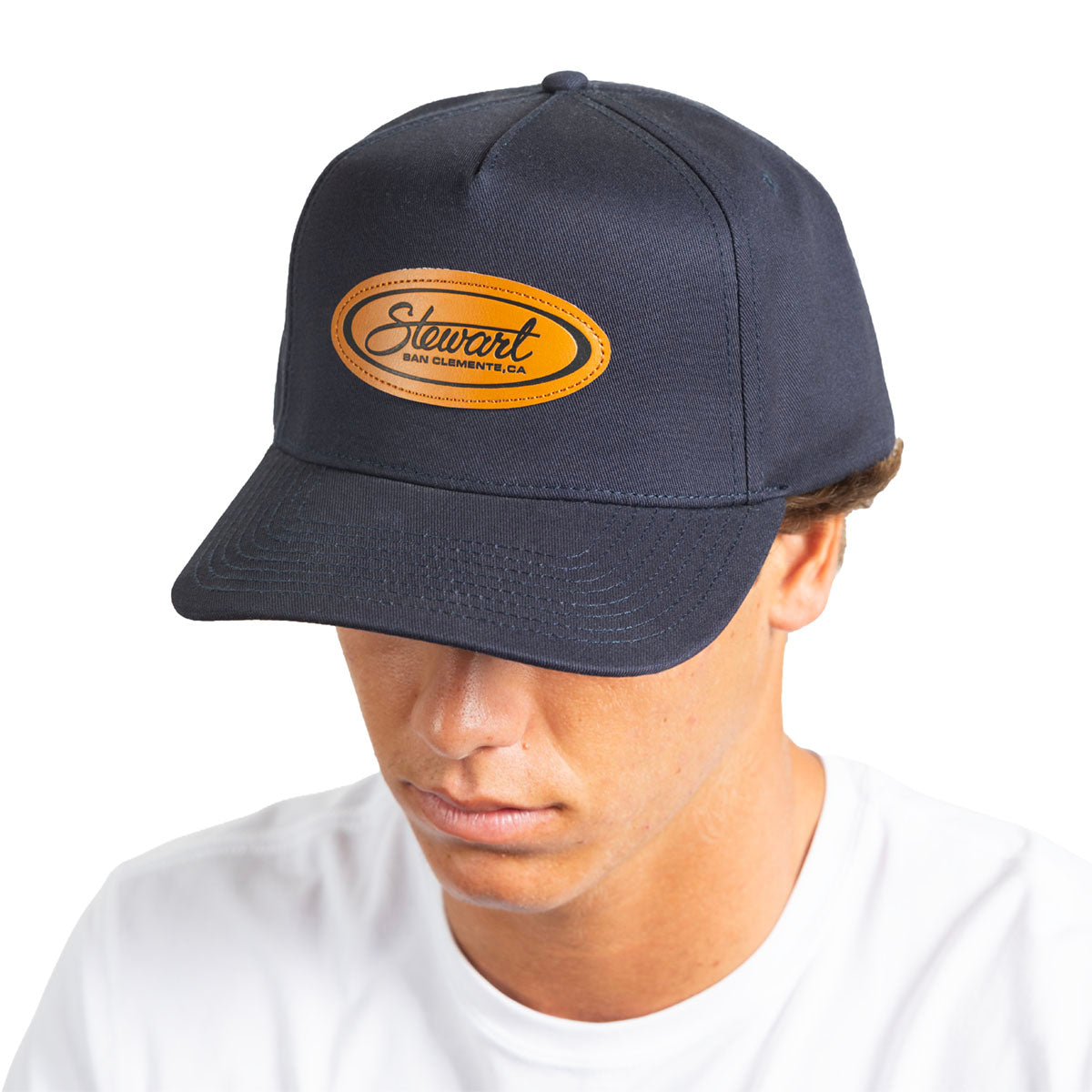 A man wearing a dark navy blue baseball cap with an oval tan leather patch on the front that reads 'Stewart San Clemente' in black script, against a plain white background.