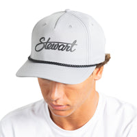 A man wearing a light gray snapback cap with the name 'Stewart' embroidered in black script on the front, above a black braided band, photographed against a plain white background.
