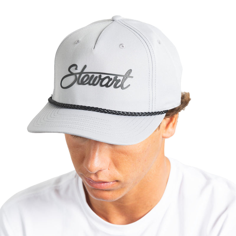 A man wearing a light gray snapback cap with the name 'Stewart' embroidered in black script on the front, above a black braided band, photographed against a plain white background.