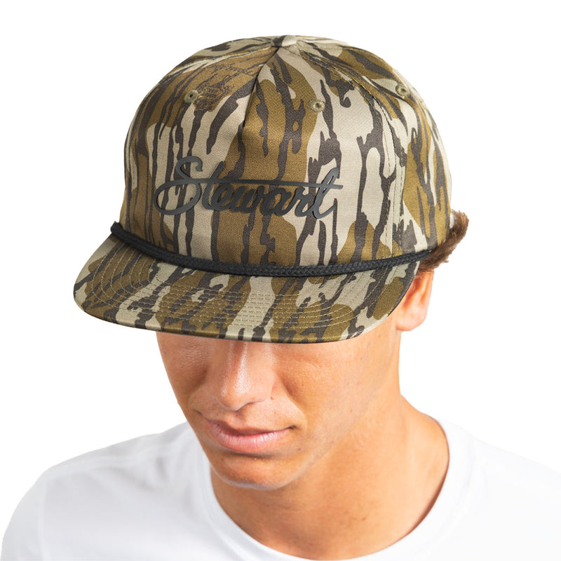 A man wearing a camouflage baseball cap with a black embroidered script logo on the front that reads 'Stewart'. The cap features a tan, brown, and olive green camo pattern with a black rope detail around the brim.