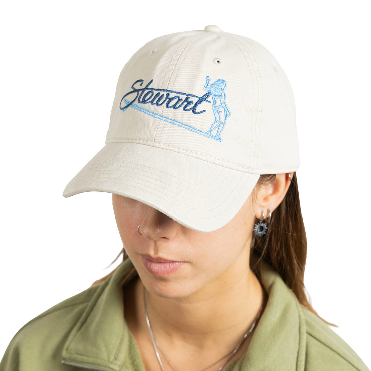 A woman wearing a light beige baseball cap with blue embroidered script reading 'Stewart' and a blue line-art figure, shown against a plain white background.