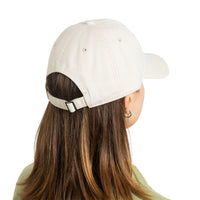 A person shown from the back wearing a light cream baseball cap with an adjustable strap and silver buckle, photographed against a plain white background.
