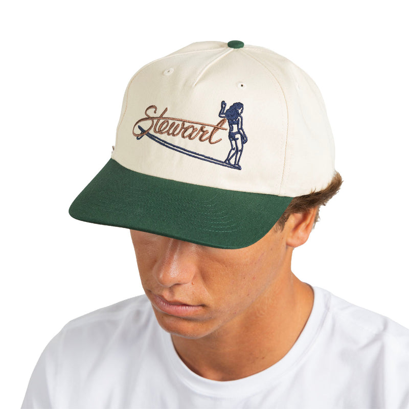 A man wearing a structured baseball cap with an off-white crown and a dark green visor. The front of the cap features a navy blue graphic of a surfer and the word 'Stewart' in brown script.