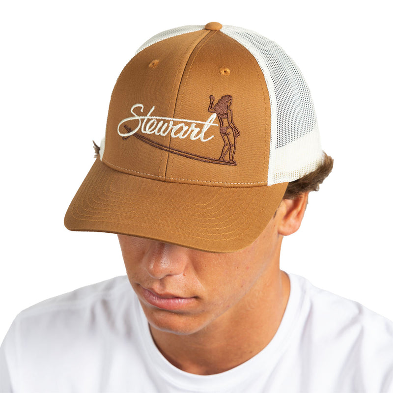A man wearing a brown and white trucker hat with 'Stewart' and a surfer graphic embroidered on the front, photographed against a plain white background.