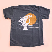 Black t-shirt with a graphic of a hand holding a surfboard against an orange sun on a peach background