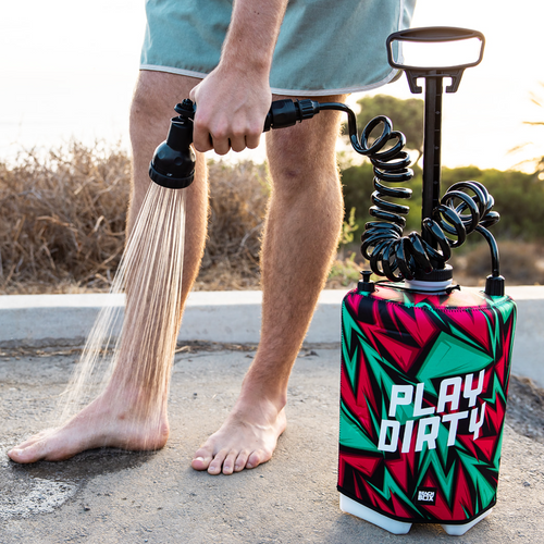 Person using a portable shower with a colorful design and 'Play Dirty' text outdoors.
