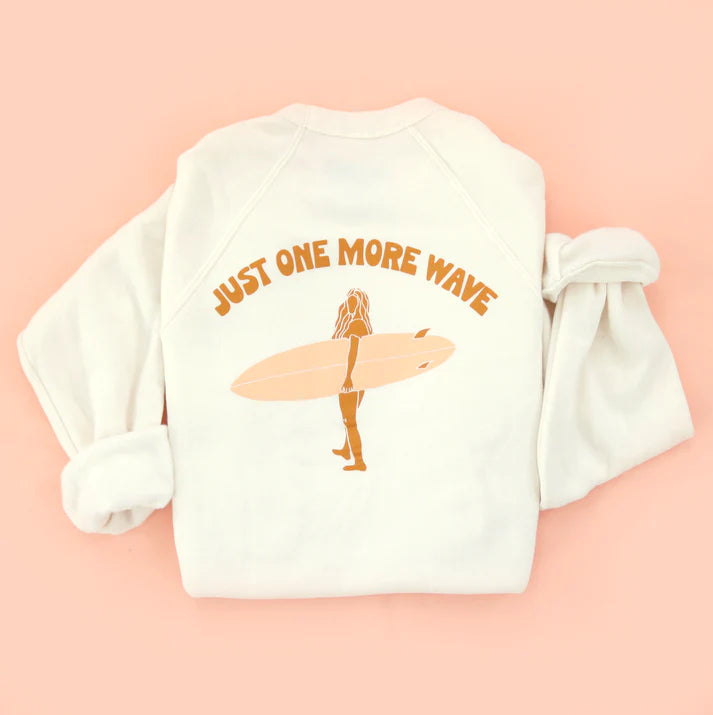 White sweatshirt with 'Just One More Wave' text and a graphic of a surfer on a pink background