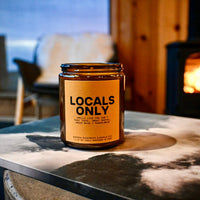Candle labeled 'Locals Only' on a stone surface with a fireplace in the background