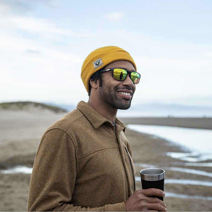 A person wearing the SUNCLOUD RAMBLER SUNGLASSES with a golden yellow beanie on a beach-like setting. The sunglasses have reflective lenses and metal frame with a logo plaque.