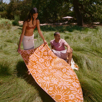 Two people with a patterned orange and white towel in a grassy outdoor setting