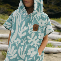 Child wearing a teal and white patterned hooded towel with a brand logo, standing outdoors.
